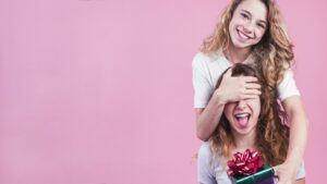 Female covering her friend s eyes giving gift box against pink background 1 2